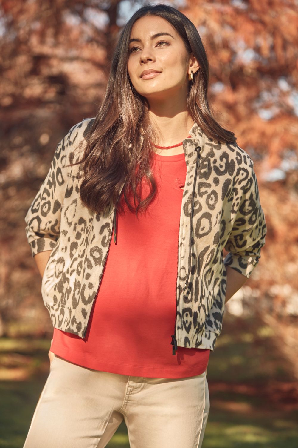 Woman wearing a red top and leopard print jacket outdoors with autumn foliage.