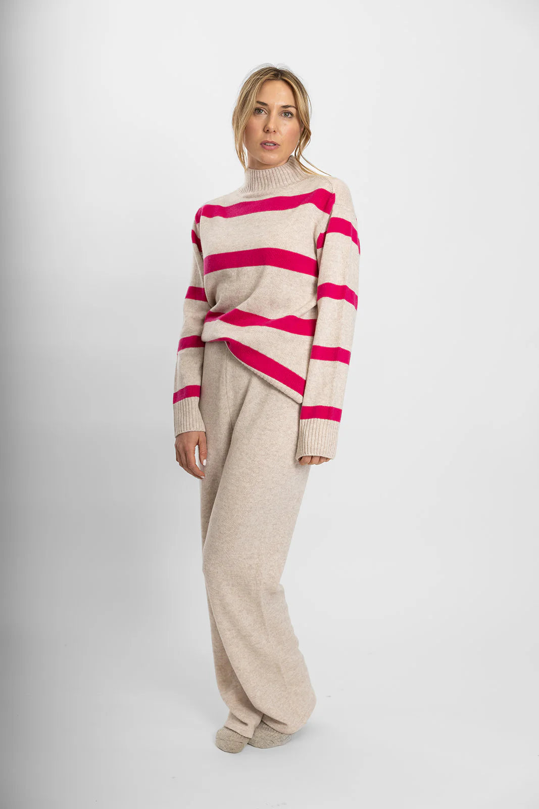A model wearing an oatmeal and bright pink striped polo neck jumper with long sleeves, paired with light-colored trousers.